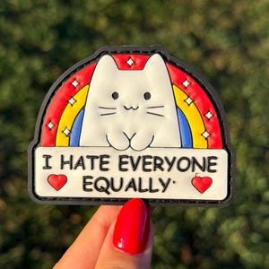 May include: A white cat peeking over a rainbow with the text "I HATE EVERYONE EQUALLY" on a black background. Two red hearts flank the text. The image is held by a hand with red nail polish.