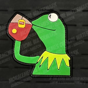 May include: Green frog puppet holding a red teacup with a yellow tea bag and the word "Lipton" printed on it. The frog is wearing a yellow collar. The background is black.
