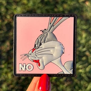 May include: A square patch featuring Bugs Bunny with a gray and white design on a pink background. The word "NO" is printed in white. The patch has a black border and is held by a hand with a red nail.