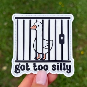 May include: A white sticker featuring a cartoon goose inside a black and white barred cage. The goose has an orange beak and feet. The text "got too silly" is below the cage in black letters.