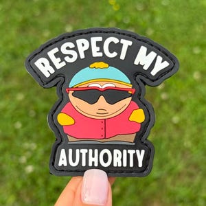 May include: A black patch with the phrase "RESPECT MY AUTHORITY" in white lettering. The patch features a cartoon character wearing sunglasses, a red jacket, and a blue hat. The background is green.
