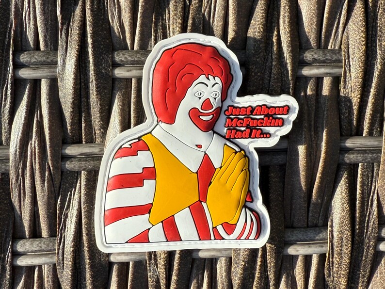 K&ouml;nnte beinhalten: Ein wei&szlig;er Patch mit einer roten und gelben Comicfigur mit roten Haaren und einem wei&szlig;-rot gestreiften Hemd. Die Figur h&auml;lt ihre H&auml;nde in einer betenden Geste zusammen. Der Text "Just About McFuckin Had It..." ist in Wei&szlig; auf schwarzem Hintergrund geschrieben.