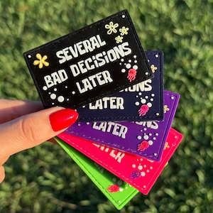 Peut inclure: Plusieurs écussons colorés sont tenus par une main. Ils affichent le texte "SEVERAL BAD DECISIONS LATER". Les écussons sont noirs, marine, violets, roses et verts, avec du texte blanc.