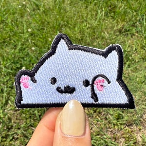 May include: Embroidered patch featuring a light blue cat design with black outlines. The cat has pink accents on its paws and eye, with a black nose and mouth. The patch is held against a background of green grass.