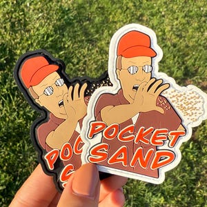 May include: Two white and black embroidered patches featuring a cartoon man wearing a red hat and sunglasses, with the text "Pocket Sand" in red letters.