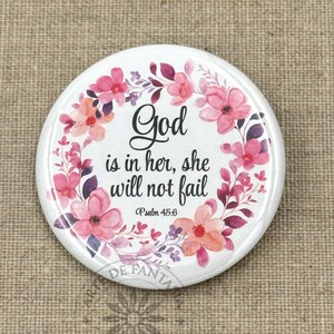 May include: A white round button with a floral wreath design. The button features the text "God is in her, she will not fail" in black script, with "Psalm 45:6" below. The floral wreath includes pink and purple flowers.