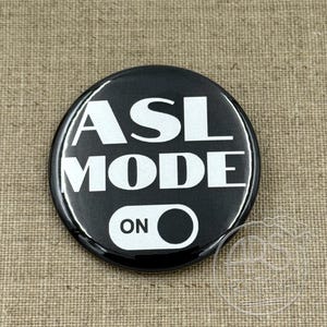 May include: A black round button with the words "ASL MODE ON" in white block letters. The button has a toggle graphic with the word "ON" in white. The button is on a tan fabric background.