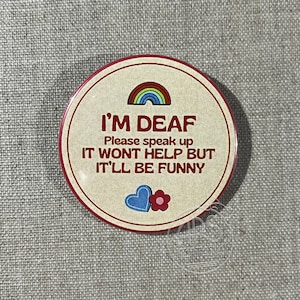 May include: A round button with a beige background and red border. The button has a rainbow, two hearts, and the text "I'M DEAF Please speak up IT WONT HELP BUT IT'LL BE FUNNY".