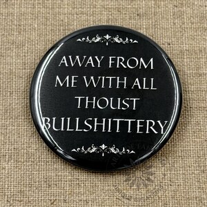 May include: A black, round button with white text that reads "AWAY FROM ME WITH ALL THOUST BULLSHITTERY." The button has a glossy finish and decorative flourishes at the top and bottom. The background is a textured, tan fabric.