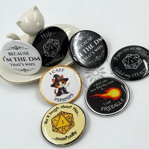 May include: A collection of round, colorful buttons with various text and graphics. The buttons feature phrases like "Because I'm the DM, that's why" and "I cast Fireball!" along with dice and cat illustrations. Some buttons are black, white, and gold.