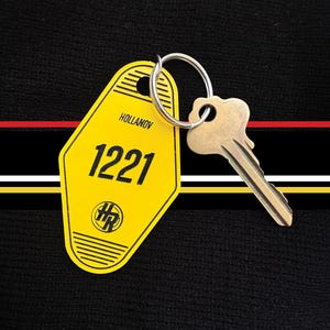 May include: A vintage-style key and yellow room key tag with the number 1221. The tag has the word "Hollanov" at the top and a logo at the bottom. The key is attached to the tag with a silver ring.