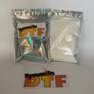Op de afbeelding: Twee rechthoekige zakjes met ritssluiting, &eacute;&eacute;n met een holografische afwerking en een wit label met de tekst Dynamite DTF, en de andere een zilverkleurig zakje met wit poeder. Een losse sticker met hetzelfde logo is inbegrepen.
