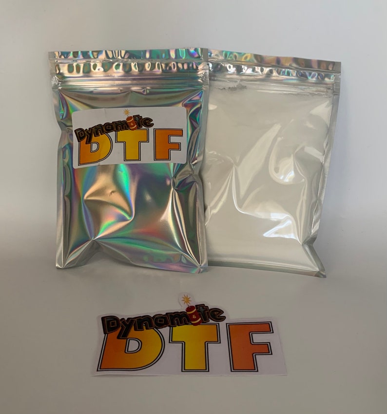 Op de afbeelding: Twee rechthoekige zakjes met ritssluiting, &eacute;&eacute;n met een holografische afwerking en een wit label met de tekst Dynamite DTF, en de andere een zilverkleurig zakje met wit poeder. Een losse sticker met hetzelfde logo is inbegrepen.