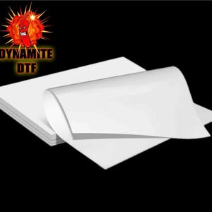 May include: A stack of white DTF transfer paper. The paper is folded in half and the top sheet is curled up. The paper is labeled "Dynamite DTF".
