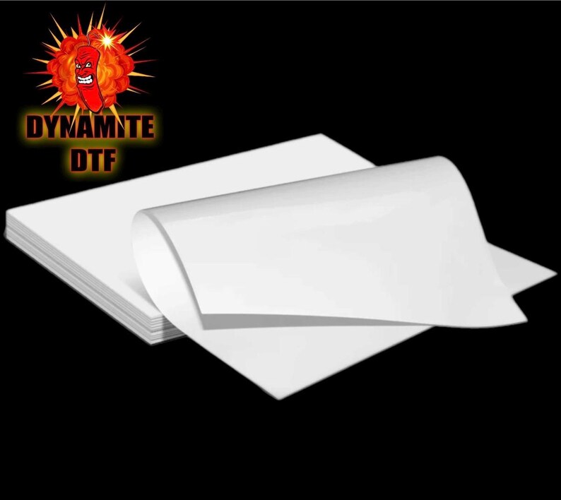 May include: A stack of white DTF transfer paper. The paper is folded in half and the top sheet is curled up. The paper is labeled "Dynamite DTF".
