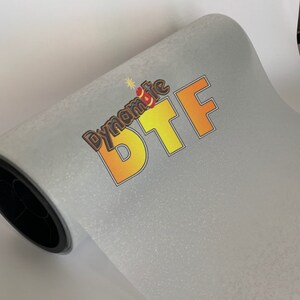 May include: A roll of white glitter vinyl with the text "Dynamite BTF" printed on it in orange and yellow letters.