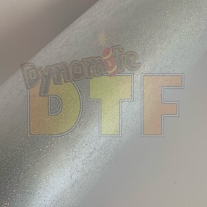 May include: A close-up of a white, glittery surface with the text "Dynamite DTF" printed in yellow and orange.