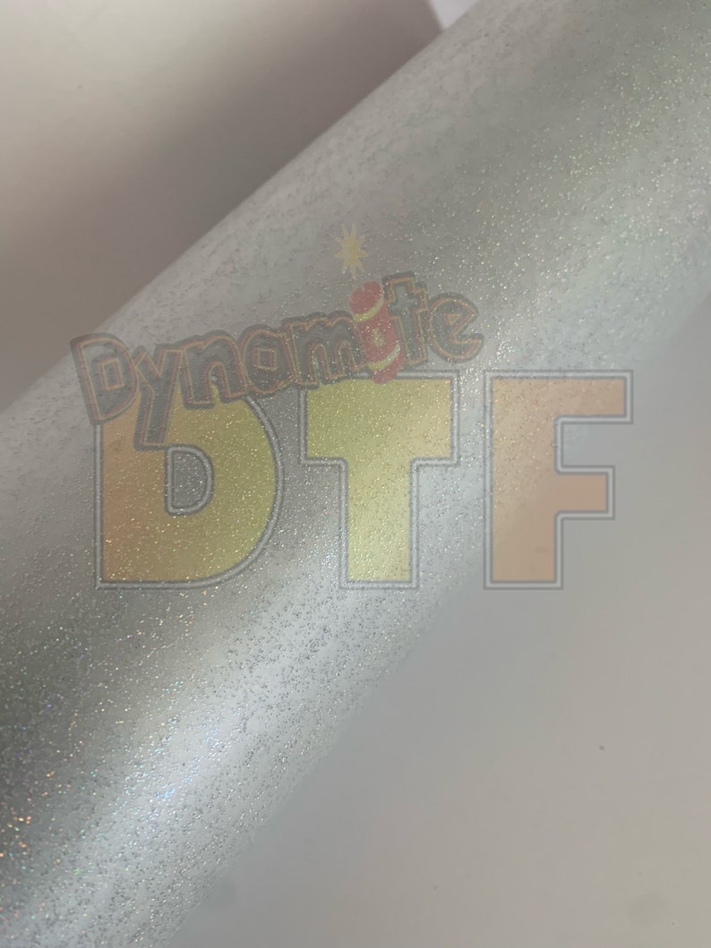 May include: A close-up of a white, glittery surface with the text "Dynamite DTF" printed in yellow and orange.