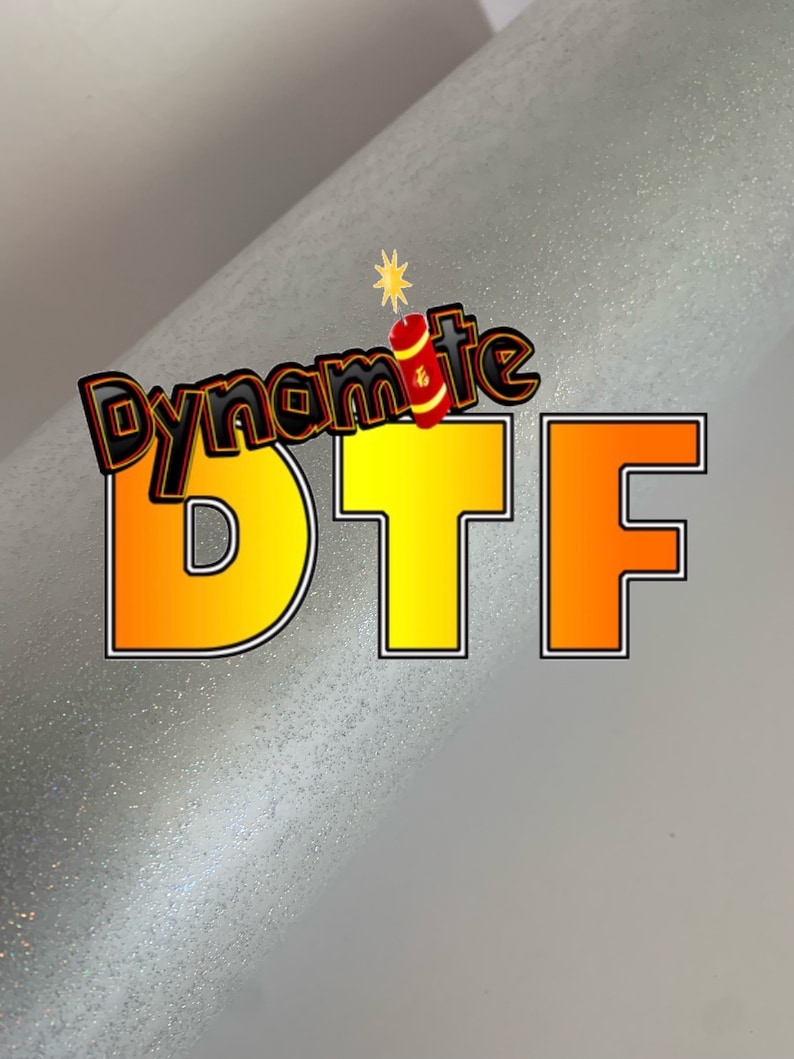 May include: A silver glitter background with the text "Dynamite DTF" in yellow and orange letters. The text is outlined in black.