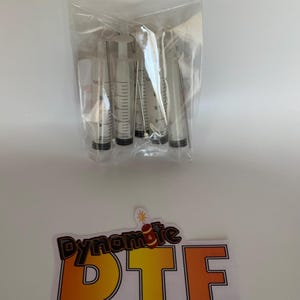 Op de afbeelding: Een doorzichtige plastic zak met vijf transparante spuiten met zwarte rubberen plunjers en metalen naalden, vergezeld van een rechthoekige sticker met de tekst Dynamite DTF en een kleine dynamiet-afbeelding boven de letters.