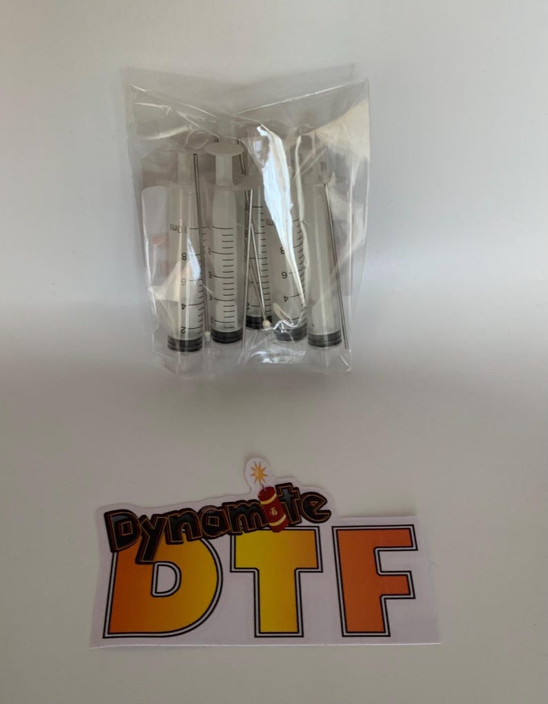 Op de afbeelding: Een doorzichtige plastic zak met vijf transparante spuiten met zwarte rubberen plunjers en metalen naalden, vergezeld van een rechthoekige sticker met de tekst Dynamite DTF en een kleine dynamiet-afbeelding boven de letters.