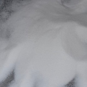 May include: A pile of fine, white, granular material with a crystalline texture. The substance consists of numerous tiny, translucent particles that reflect light, creating a soft, uniform appearance across the surface of the mound.