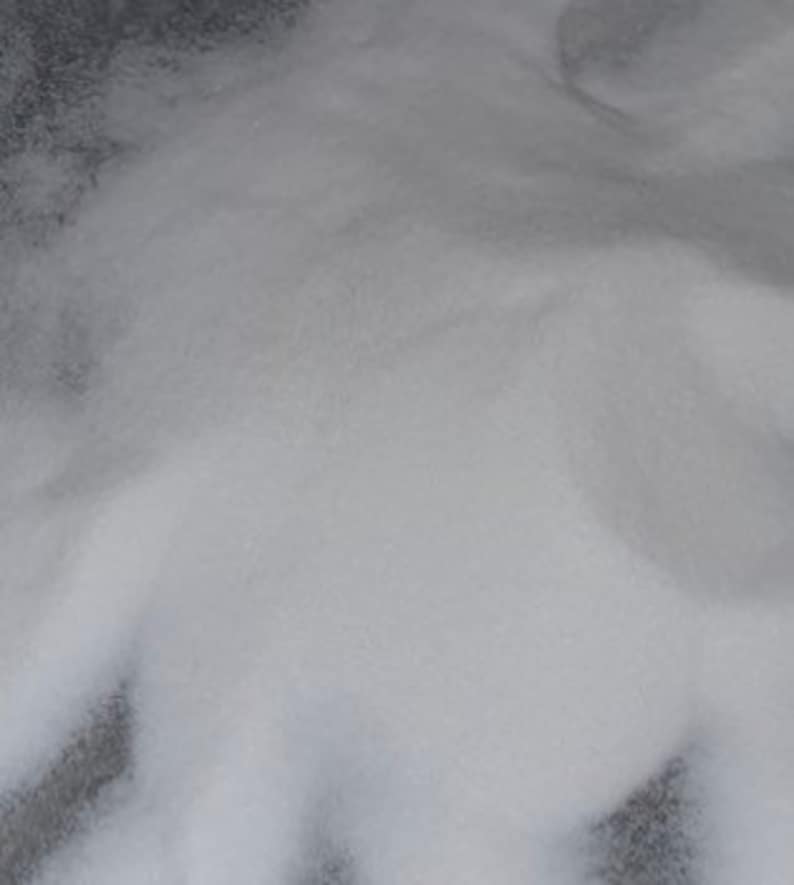 May include: A pile of fine, white, granular material with a crystalline texture. The substance consists of numerous tiny, translucent particles that reflect light, creating a soft, uniform appearance across the surface of the mound.