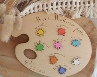 Puede incluir: Paleta de artista de madera con salpicaduras de pintura de colores y las palabras "Merci Maîtresse pour" grabadas. La paleta incluye el nombre "Giulia" y el año "2022-2023". Artículo decorativo con temática de agradecimiento a profesores.