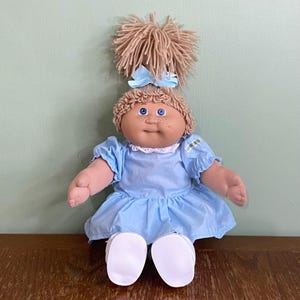 May include: A doll with tan skin, blue eyes, and freckles, featuring light brown yarn hair styled in a high ponytail with a light blue bow. The doll wears a light blue short-sleeved dress with a white eyelet collar and plain white shoes.