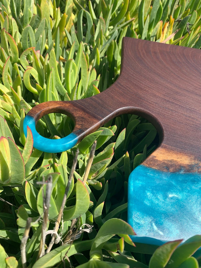 May include: A wooden cutting board with a blue resin inlay and a handle. The board is made of walnut wood and has a natural finish. The resin inlay is a light blue color and has a swirling pattern.