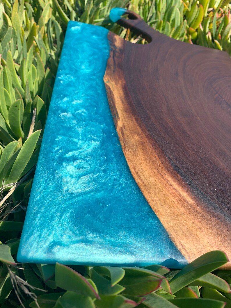 May include: A large wooden cutting board with a blue epoxy resin inlay. The resin has a swirling pattern and a glittery finish. The board has a handle on one end.