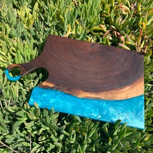 May include: A large wooden cutting board with a blue epoxy resin inlay. The board has a handle and is made of dark wood.