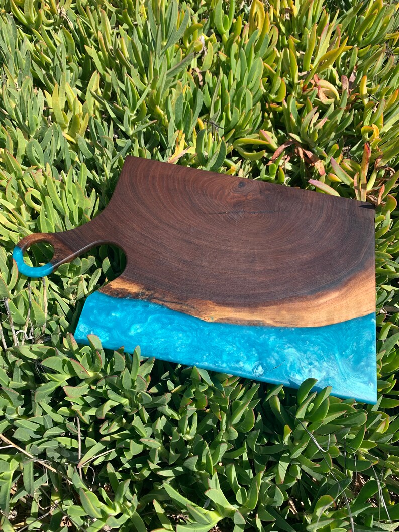 May include: A large wooden cutting board with a blue epoxy resin inlay. The board has a handle and is made of dark wood.