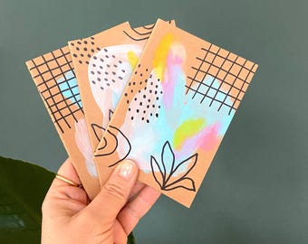May include: Three kraft paper greeting cards with abstract watercolour designs in pastel colours. Each card features unique patterns, including black grids, dots, and floral outlines. The cards are held in a hand against a dark green background.