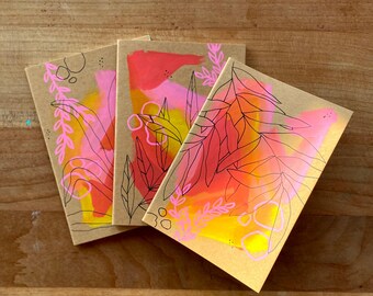 May include: Three notebooks with abstract art on kraft paper. Each has a unique design with vibrant watercolour splashes of pink, orange, and yellow, accented by black and pink line drawings of leaves and floral elements.
