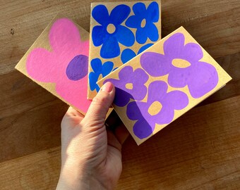 May include: Three floral-themed greeting cards. One card features a large pink flower with a purple centre. The other two cards have blue and purple flowers with yellow centres. The cards are on a brown background.
