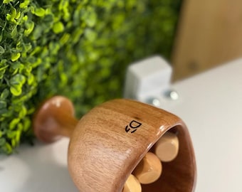 Könnte beinhalten: Ein Massage-Werkzeug aus Holz mit einer abgerundeten Becherform und einem Griff. Der Becher enthält drei kleine, hellfarbene Holzkugeln. Das Werkzeug hat eine glatte, polierte Oberfläche und eine dunkle Holzmaserung.