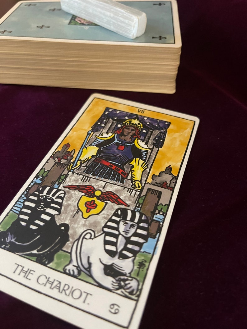 May include: The Chariot tarot card features a colorful illustration of a charioteer in armor, driving a chariot pulled by two sphinxes. The card is set against a backdrop of a starry night sky and a cityscape. The card number VII is displayed in the top right corner.