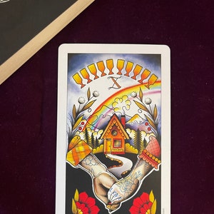 May include: Tarot card depicting the Ten of Cups, featuring a rainbow, a small house, and two hands holding each other. The hands are tattooed with hearts and the words "I love" and "you".