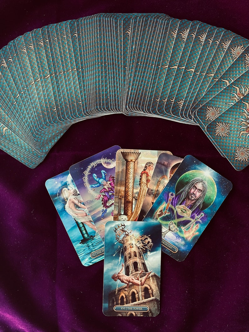 Peut inclure: Un jeu de tarot avec un design bleu et or. Les cartes sont &eacute;tal&eacute;es en demi-cercle. Cinq cartes sont dispos&eacute;es devant le jeu. Les cartes montrent des illustrations color&eacute;es de personnes et de sc&egrave;nes. Les cartes sont &eacute;tiquet&eacute;es avec des chiffres romains et les noms des cartes. Les cartes sont : Le Mat, La Tour, Le Magicien, L'&Eacute;toile et Le Chariot.