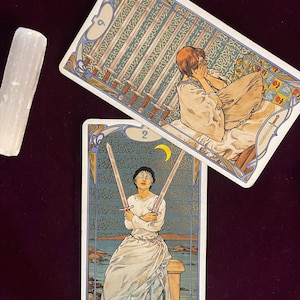 May include: Two tarot cards, the Nine of Swords and the Two of Swords. The Nine of Swords card depicts a woman with her head in her hands, surrounded by nine swords. The Two of Swords card shows a woman blindfolded, holding two swords crossed in front of her.