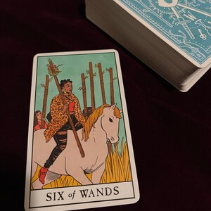 Peut inclure: Une carte de tarot avec un fond bleu clair et une bordure blanche. La carte repr&eacute;sente une femme chevauchant un cheval blanc &agrave; travers un champ d'herbe haute. La femme porte une tenue l&eacute;opard et une chemise rouge. La carte est intitul&eacute;e "Six de B&acirc;tons".