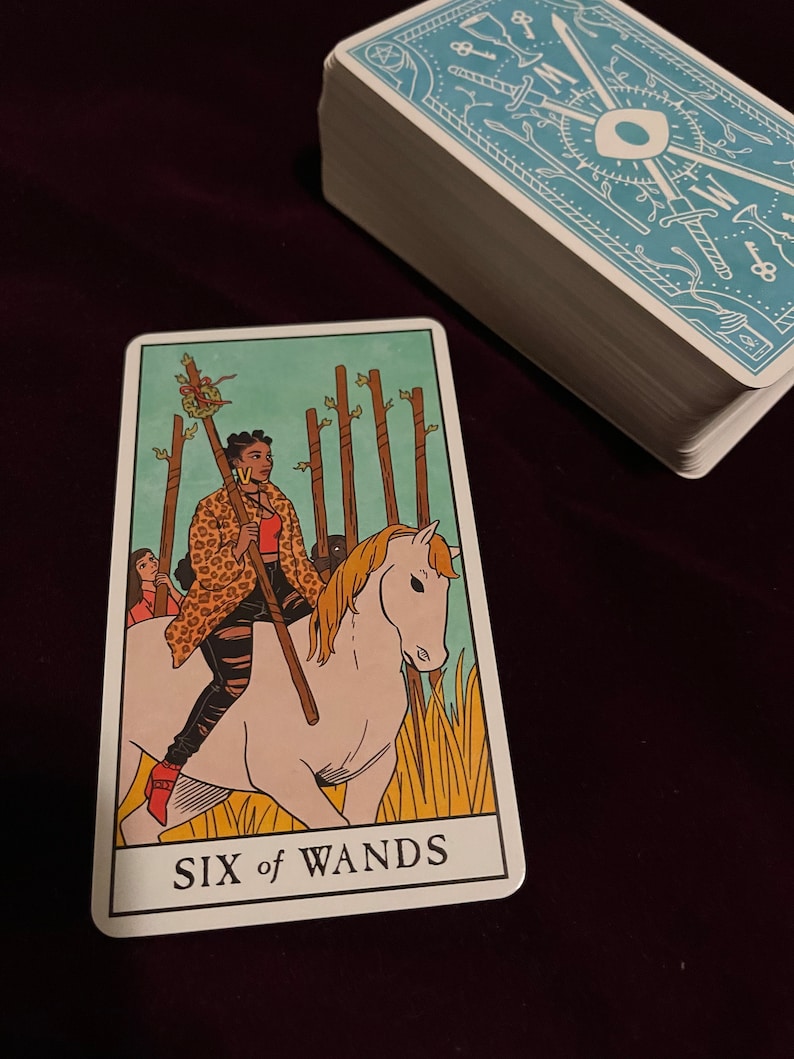 May include: A tarot card depicting a woman riding a white horse, holding a staff. The card is blue and green with the text "SIX of WANDS" at the bottom.