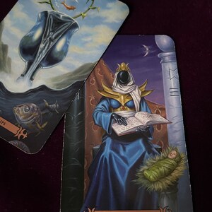 May include: Tarot card depicting the High Priestess, the second card in the Major Arcana. The card features a robed figure sitting on a throne, holding a book and a baby. The background is a dark blue with a crescent moon and stars.