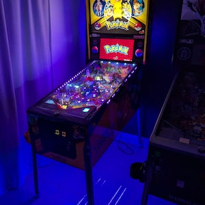 Puede incluir: Una máquina de pinball con temática de Pokemon que presenta un panel trasero iluminado con ilustraciones de personajes, una pantalla digital con el logo de Pokemon y un campo de juego con luces LED moradas y blancas sobre patas de metal.