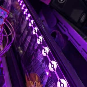 Könnte beinhalten: Eine Flipper-Seitenleiste mit einer Reihe beleuchteter violetter LED-Lichter in Form von kreisförmigen Poké-Bällen. Das dunkle Gehäuse zeigt eine Grafik eines braunen, stacheligen Wesens mit Krallen, und links ist eine Drahtform aus Metall zu sehen.