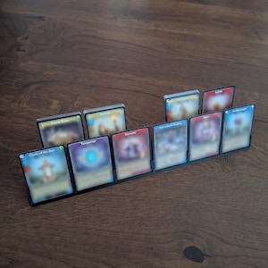 May include: A black plastic card holder displaying several trading cards. The cards have various colorful designs and text, with titles like "Cleric of the Sun" and "Teleporter". The holder is on a wooden surface.