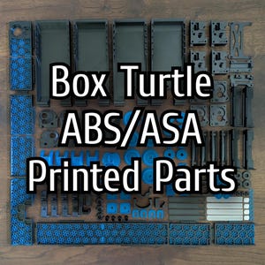 May include: A collection of black and blue 3D-printed components, featuring various shapes, laid out on a wooden background. The image includes the text "Box Turtle ABS/ASA Printed Parts".