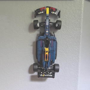 Könnte beinhalten: Ein detailliertes Modell eines Formel-1-Rennwagens, überwiegend marineblau mit roten und gelben Akzenten. Das Modell zeigt Sponsorenlogos und ist an einer hellgrauen Wand befestigt. Die Räder sind schwarz.