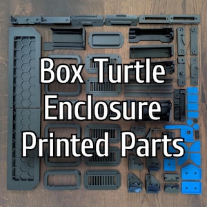Może przedstawiać: Zestaw ciemnoszarych i niebieskich części drukowanych w 3D do obudowy żółwia pudełkowego, ułożonych na drewnianej powierzchni. Części obejmują różne kształty i rozmiary, takie jak prostokątne ramy, otwory wentylacyjne i panele. Widoczny jest tekst "Box Turtle Enclosure Printed Parts".